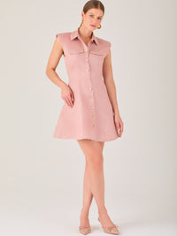 Este vestido sin mangas tiene un diseño camisero de corte recto con estructura entallada en la parte superior que se ajusta a la silueta. Tiene cuello tipo camisa y una botonadura completa al frente con botones grandes y visibles. A la altura del pecho, incluye dos bolsillos de parche con solapa, añadiendo un detalle funcional y decorativo. La falda tiene una caída en línea A, que da un toque femenino y ligeramente acampanado. La parte trasera mantiene la sencillez con costuras que definen la forma del vest