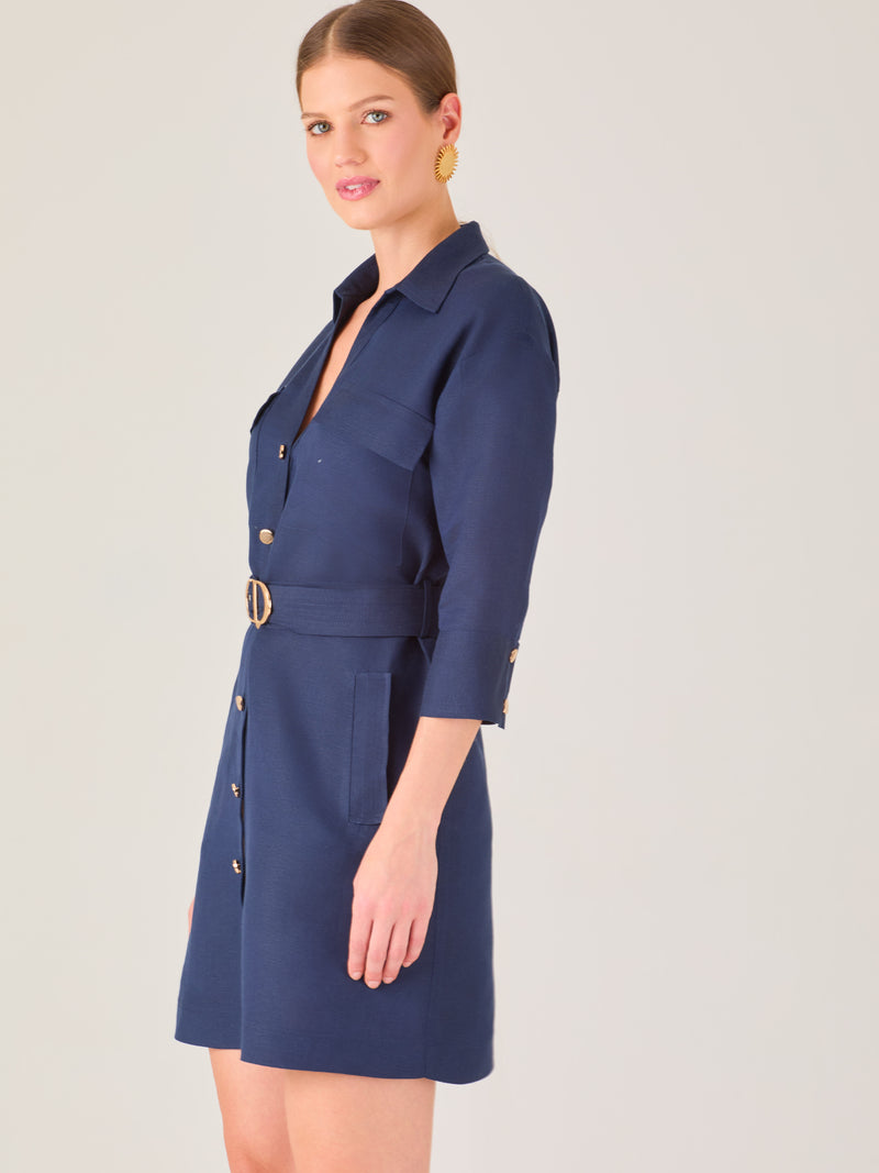 Vestido tipo camisero corto, con cuello clásico, mangas tres cuartos, botones frontales y cinturón con hebilla dorada. Estilo elegante y casual.