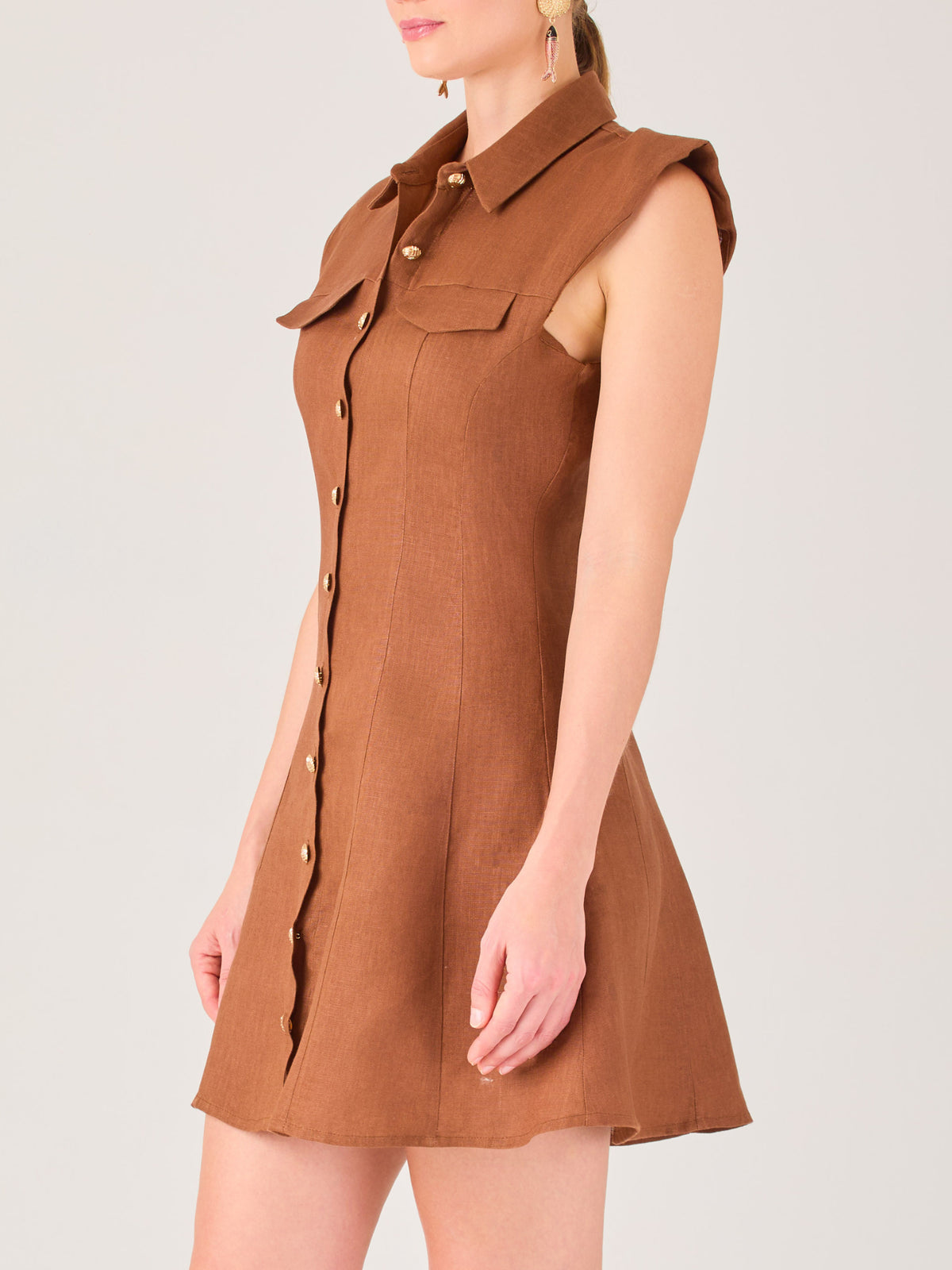 Este vestido sin mangas tiene un diseño camisero de corte recto con estructura entallada en la parte superior que se ajusta a la silueta. Tiene cuello tipo camisa y una botonadura completa al frente con botones grandes y visibles. A la altura del pecho, incluye dos bolsillos de parche con solapa, añadiendo un detalle funcional y decorativo. La falda tiene una caída en línea A, que da un toque femenino y ligeramente acampanado. La parte trasera mantiene la sencillez con costuras que definen la forma del vest
