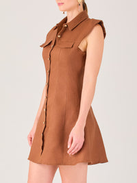 Este vestido sin mangas tiene un diseño camisero de corte recto con estructura entallada en la parte superior que se ajusta a la silueta. Tiene cuello tipo camisa y una botonadura completa al frente con botones grandes y visibles. A la altura del pecho, incluye dos bolsillos de parche con solapa, añadiendo un detalle funcional y decorativo. La falda tiene una caída en línea A, que da un toque femenino y ligeramente acampanado. La parte trasera mantiene la sencillez con costuras que definen la forma del vest
