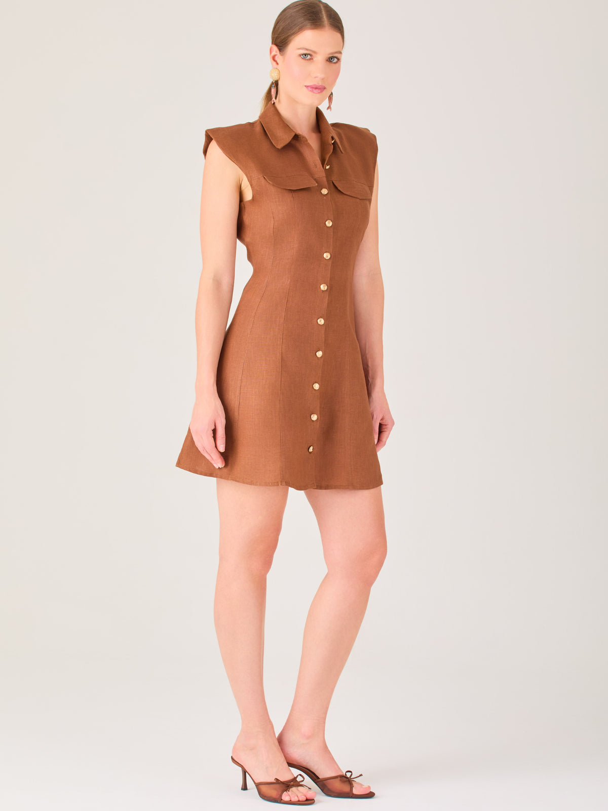 Este vestido sin mangas tiene un diseño camisero de corte recto con estructura entallada en la parte superior que se ajusta a la silueta. Tiene cuello tipo camisa y una botonadura completa al frente con botones grandes y visibles. A la altura del pecho, incluye dos bolsillos de parche con solapa, añadiendo un detalle funcional y decorativo. La falda tiene una caída en línea A, que da un toque femenino y ligeramente acampanado. La parte trasera mantiene la sencillez con costuras que definen la forma del vest