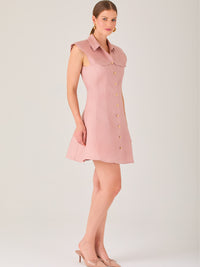 Este vestido sin mangas tiene un diseño camisero de corte recto con estructura entallada en la parte superior que se ajusta a la silueta. Tiene cuello tipo camisa y una botonadura completa al frente con botones grandes y visibles. A la altura del pecho, incluye dos bolsillos de parche con solapa, añadiendo un detalle funcional y decorativo. La falda tiene una caída en línea A, que da un toque femenino y ligeramente acampanado. La parte trasera mantiene la sencillez con costuras que definen la forma del vest