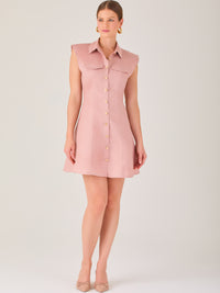 Este vestido sin mangas tiene un diseño camisero de corte recto con estructura entallada en la parte superior que se ajusta a la silueta. Tiene cuello tipo camisa y una botonadura completa al frente con botones grandes y visibles. A la altura del pecho, incluye dos bolsillos de parche con solapa, añadiendo un detalle funcional y decorativo. La falda tiene una caída en línea A, que da un toque femenino y ligeramente acampanado. La parte trasera mantiene la sencillez con costuras que definen la forma del vest