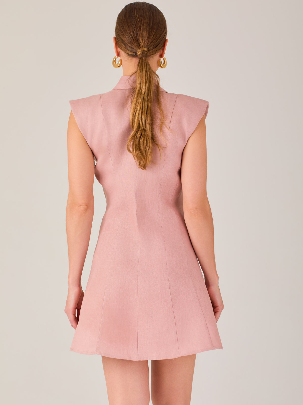 Este vestido sin mangas tiene un diseño camisero de corte recto con estructura entallada en la parte superior que se ajusta a la silueta. Tiene cuello tipo camisa y una botonadura completa al frente con botones grandes y visibles. A la altura del pecho, incluye dos bolsillos de parche con solapa, añadiendo un detalle funcional y decorativo. La falda tiene una caída en línea A, que da un toque femenino y ligeramente acampanado. La parte trasera mantiene la sencillez con costuras que definen la forma del vest