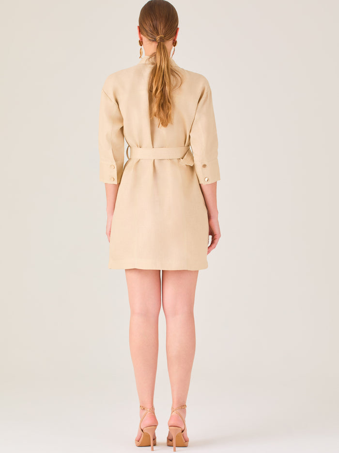Vestido tipo camisero beige, corto, con cuello clásico, mangas tres cuartos, botones frontales y cinturón con hebilla dorada. Estilo elegante y casual.