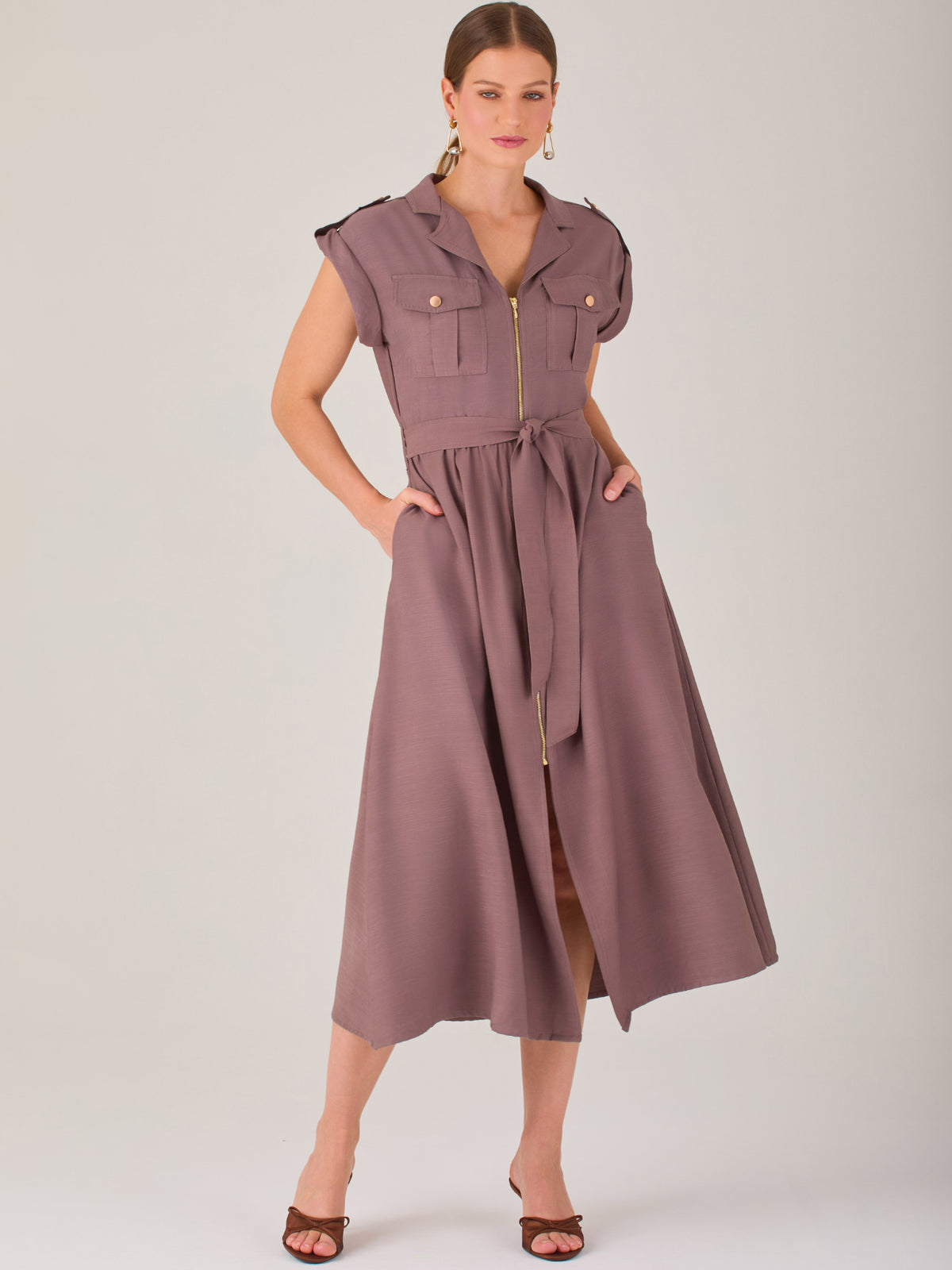 Diseño atemporal con carácter. Este vestido midi sin mangas combina estructura y feminidad con detalles utilitarios como bolsillos frontales, cierre metálico y solapas marcadas. El cinturón en la cintura permite ajustar la silueta, creando un look pulido ideal para la oficina, eventos casuales o estilismos urbanos con elegancia natural.
