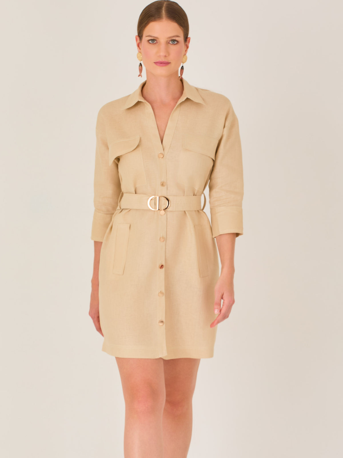 Vestido tipo camisero beige, corto, con cuello clásico, mangas tres cuartos, botones frontales y cinturón con hebilla dorada. Estilo elegante y casual.