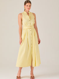 Este vestido es de corte midi, sin mangas y en un tono amarillo suave. Presenta un diseño tipo camisero con botones al frente que recorren toda la longitud del vestido. El cuello es tipo mao, lo que le da un aire moderno y sofisticado. Tiene una cinturilla marcada con un cinturón del mismo tejido y color que se ajusta con hebilla metálica, aportando estructura y definiendo la silueta. La falda cae con una ligera amplitud gracias a los pliegues sutiles que nacen desde la cintura, ofreciendo movimiento y fres