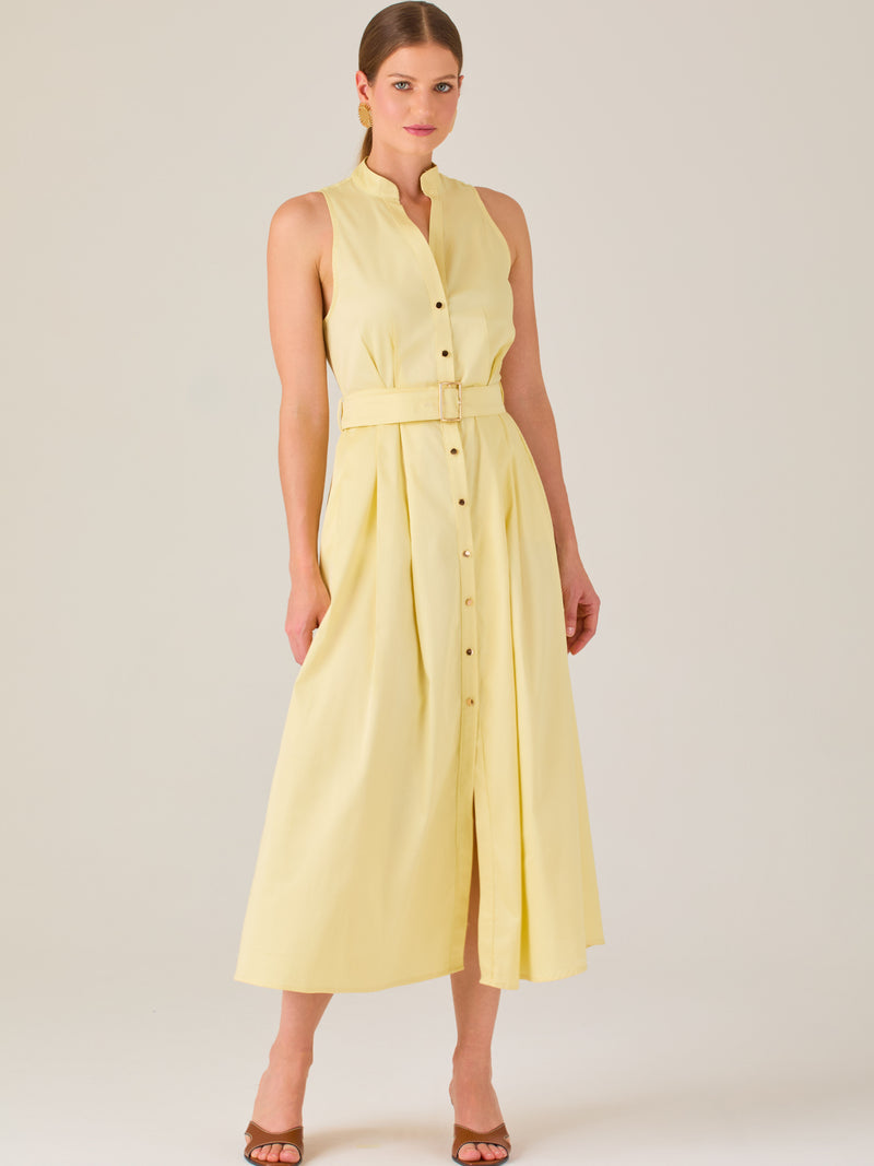 Este vestido es de corte midi, sin mangas y en un tono amarillo suave. Presenta un diseño tipo camisero con botones al frente que recorren toda la longitud del vestido. El cuello es tipo mao, lo que le da un aire moderno y sofisticado. Tiene una cinturilla marcada con un cinturón del mismo tejido y color que se ajusta con hebilla metálica, aportando estructura y definiendo la silueta. La falda cae con una ligera amplitud gracias a los pliegues sutiles que nacen desde la cintura, ofreciendo movimiento y fres