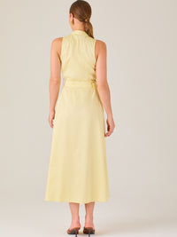 Este vestido es de corte midi, sin mangas y en un tono amarillo suave. Presenta un diseño tipo camisero con botones al frente que recorren toda la longitud del vestido. El cuello es tipo mao, lo que le da un aire moderno y sofisticado. Tiene una cinturilla marcada con un cinturón del mismo tejido y color que se ajusta con hebilla metálica, aportando estructura y definiendo la silueta. La falda cae con una ligera amplitud gracias a los pliegues sutiles que nacen desde la cintura, ofreciendo movimiento y fres