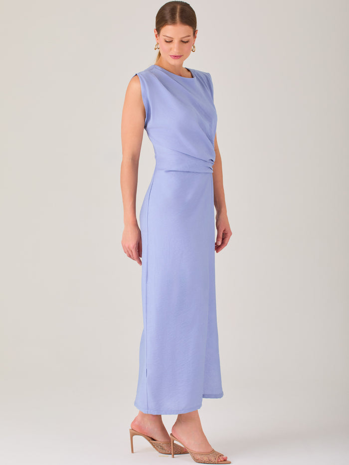 Este vestido es largo y sin mangas, con un diseño elegante y minimalista. Tiene un escote cerrado redondo y un detalle drapeado en la parte frontal del torso, que crea una ligera asimetría visual y acentúa la silueta. Este drapeado se sostiene con una hebilla decorativa al costado de la cintura, lo que añade un toque sofisticado y estilizado. La falda cae de manera recta hasta los tobillos, con una pequeña abertura lateral que proporciona comodidad y un leve movimiento al caminar. El corte general es limpio