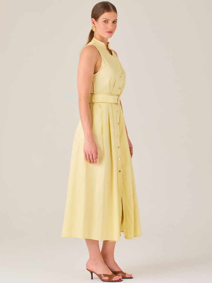 Este vestido es de corte midi, sin mangas y en un tono amarillo suave. Presenta un diseño tipo camisero con botones al frente que recorren toda la longitud del vestido. El cuello es tipo mao, lo que le da un aire moderno y sofisticado. Tiene una cinturilla marcada con un cinturón del mismo tejido y color que se ajusta con hebilla metálica, aportando estructura y definiendo la silueta. La falda cae con una ligera amplitud gracias a los pliegues sutiles que nacen desde la cintura, ofreciendo movimiento y fres