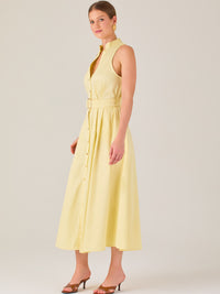 Este vestido es de corte midi, sin mangas y en un tono amarillo suave. Presenta un diseño tipo camisero con botones al frente que recorren toda la longitud del vestido. El cuello es tipo mao, lo que le da un aire moderno y sofisticado. Tiene una cinturilla marcada con un cinturón del mismo tejido y color que se ajusta con hebilla metálica, aportando estructura y definiendo la silueta. La falda cae con una ligera amplitud gracias a los pliegues sutiles que nacen desde la cintura, ofreciendo movimiento y fres