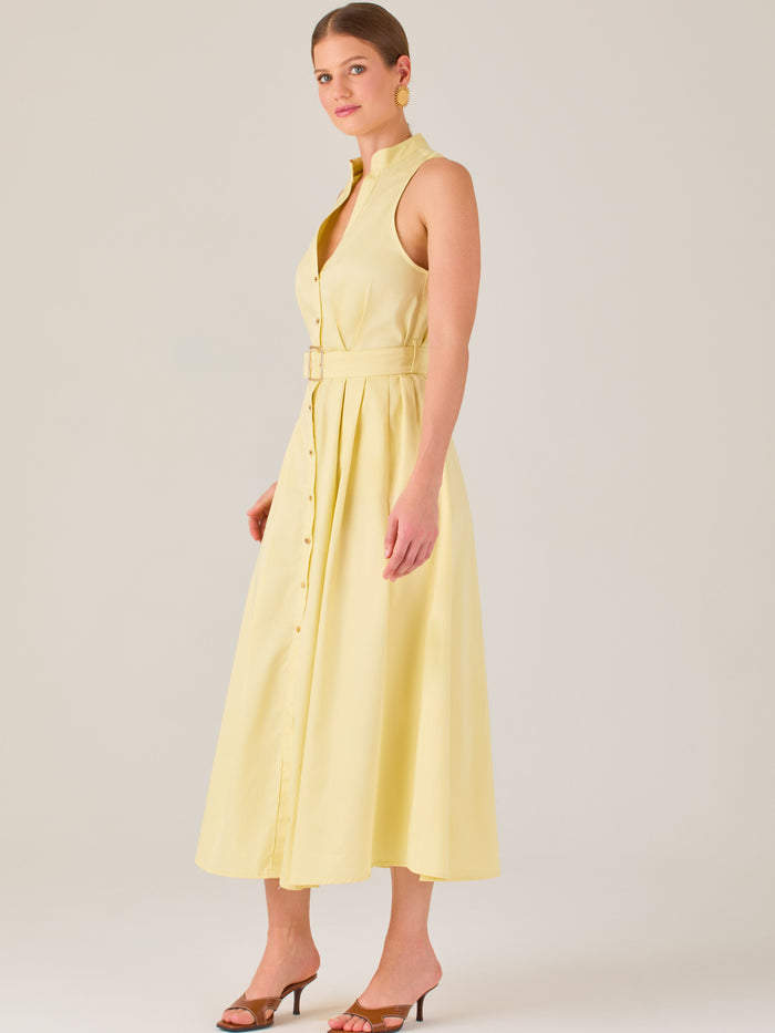 Este vestido es de corte midi, sin mangas y en un tono amarillo suave. Presenta un diseño tipo camisero con botones al frente que recorren toda la longitud del vestido. El cuello es tipo mao, lo que le da un aire moderno y sofisticado. Tiene una cinturilla marcada con un cinturón del mismo tejido y color que se ajusta con hebilla metálica, aportando estructura y definiendo la silueta. La falda cae con una ligera amplitud gracias a los pliegues sutiles que nacen desde la cintura, ofreciendo movimiento y fres