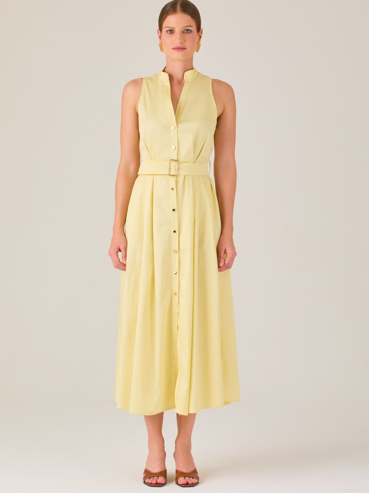 Este vestido es de corte midi, sin mangas y en un tono amarillo suave. Presenta un diseño tipo camisero con botones al frente que recorren toda la longitud del vestido. El cuello es tipo mao, lo que le da un aire moderno y sofisticado. Tiene una cinturilla marcada con un cinturón del mismo tejido y color que se ajusta con hebilla metálica, aportando estructura y definiendo la silueta. La falda cae con una ligera amplitud gracias a los pliegues sutiles que nacen desde la cintura, ofreciendo movimiento y fres