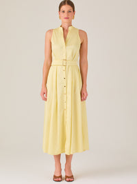 Este vestido es de corte midi, sin mangas y en un tono amarillo suave. Presenta un diseño tipo camisero con botones al frente que recorren toda la longitud del vestido. El cuello es tipo mao, lo que le da un aire moderno y sofisticado. Tiene una cinturilla marcada con un cinturón del mismo tejido y color que se ajusta con hebilla metálica, aportando estructura y definiendo la silueta. La falda cae con una ligera amplitud gracias a los pliegues sutiles que nacen desde la cintura, ofreciendo movimiento y fres