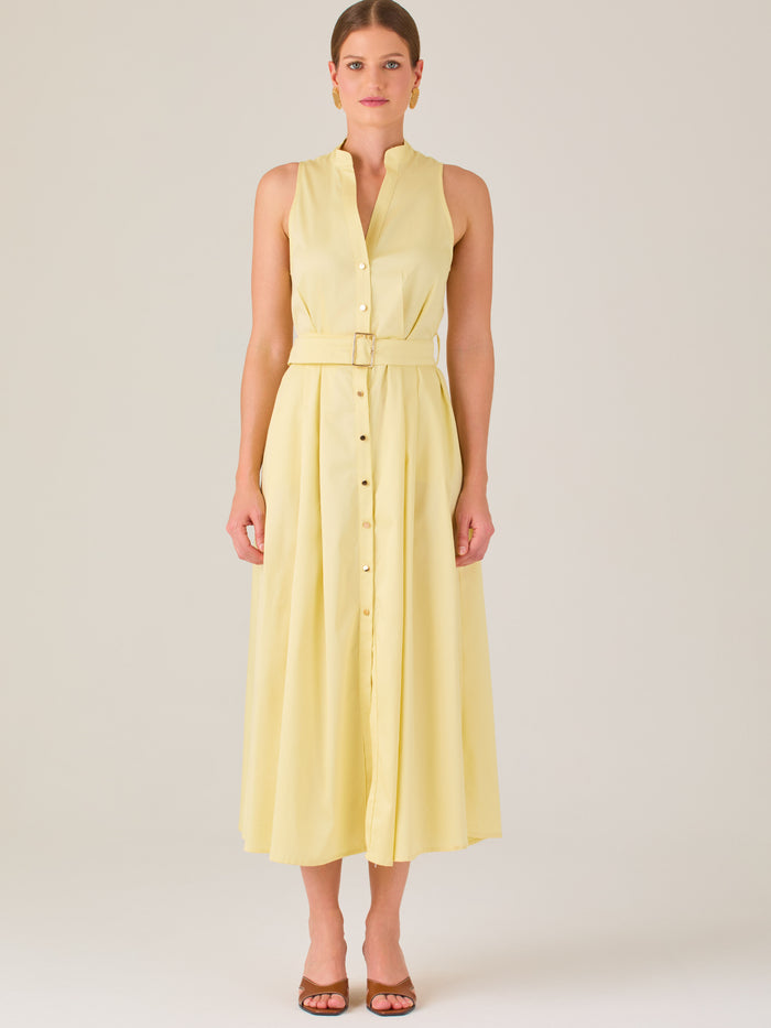 Este vestido es de corte midi, sin mangas y en un tono amarillo suave. Presenta un diseño tipo camisero con botones al frente que recorren toda la longitud del vestido. El cuello es tipo mao, lo que le da un aire moderno y sofisticado. Tiene una cinturilla marcada con un cinturón del mismo tejido y color que se ajusta con hebilla metálica, aportando estructura y definiendo la silueta. La falda cae con una ligera amplitud gracias a los pliegues sutiles que nacen desde la cintura, ofreciendo movimiento y fres