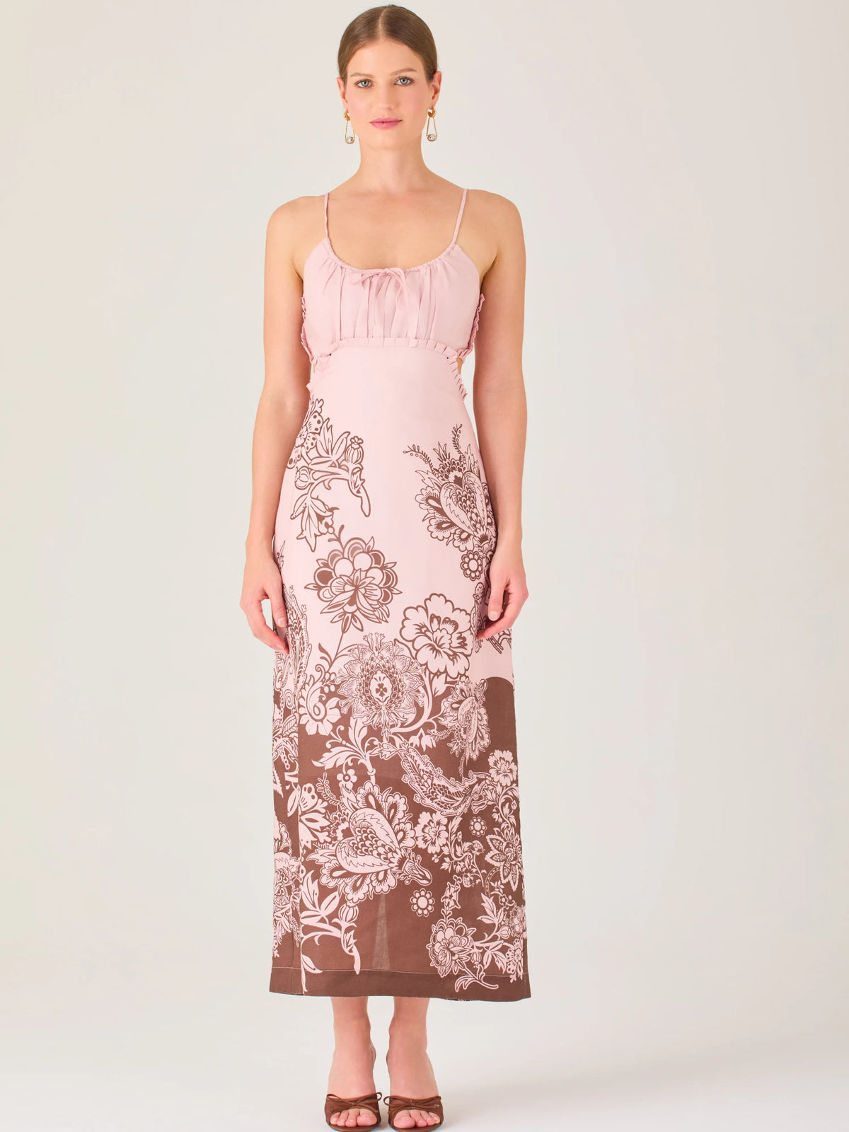 El vestido es largo y de tirantes finos, con escote fruncido y cortes laterales que dejan parte de la cintura descubierta. La falda presenta un estampado tipo floral y paisley en tonos azul profundo sobre fondo blanco, concentrado en la parte baja y difuminándose hacia arriba, creando un contraste elegante y veraniego. El diseño combina frescura y sofisticación, ideal para climas cálidos.