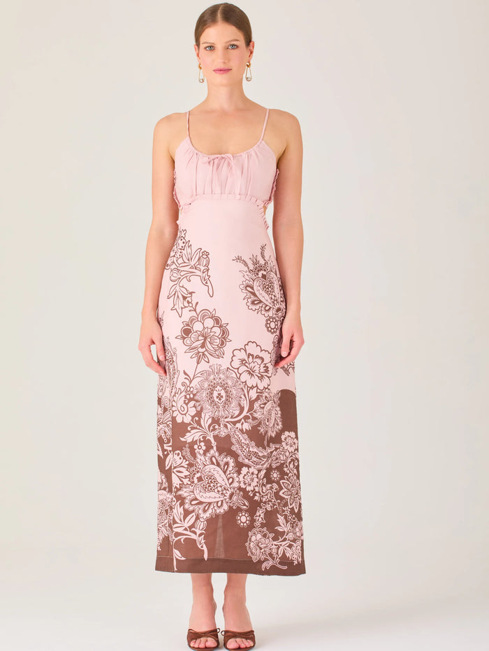 El vestido es largo y de tirantes finos, con escote fruncido y cortes laterales que dejan parte de la cintura descubierta. La falda presenta un estampado tipo floral y paisley en tonos azul profundo sobre fondo blanco, concentrado en la parte baja y difuminándose hacia arriba, creando un contraste elegante y veraniego. El diseño combina frescura y sofisticación, ideal para climas cálidos.
