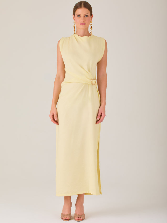 Este vestido es largo y sin mangas, con un diseño elegante y minimalista. Tiene un escote cerrado redondo y un detalle drapeado en la parte frontal del torso, que crea una ligera asimetría visual y acentúa la silueta. Este drapeado se sostiene con una hebilla decorativa al costado de la cintura, lo que añade un toque sofisticado y estilizado. La falda cae de manera recta hasta los tobillos, con una pequeña abertura lateral que proporciona comodidad y un leve movimiento al caminar. El corte general es limpio