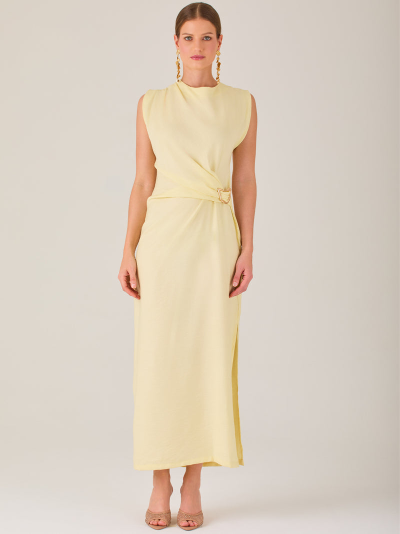 Este vestido es largo y sin mangas, con un diseño elegante y minimalista. Tiene un escote cerrado redondo y un detalle drapeado en la parte frontal del torso, que crea una ligera asimetría visual y acentúa la silueta. Este drapeado se sostiene con una hebilla decorativa al costado de la cintura, lo que añade un toque sofisticado y estilizado. La falda cae de manera recta hasta los tobillos, con una pequeña abertura lateral que proporciona comodidad y un leve movimiento al caminar. El corte general es limpio