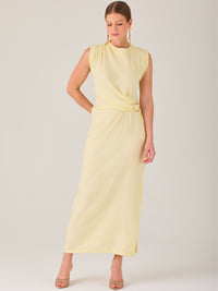 Este vestido es largo y sin mangas, con un diseño elegante y minimalista. Tiene un escote cerrado redondo y un detalle drapeado en la parte frontal del torso, que crea una ligera asimetría visual y acentúa la silueta. Este drapeado se sostiene con una hebilla decorativa al costado de la cintura, lo que añade un toque sofisticado y estilizado. La falda cae de manera recta hasta los tobillos, con una pequeña abertura lateral que proporciona comodidad y un leve movimiento al caminar. El corte general es limpio