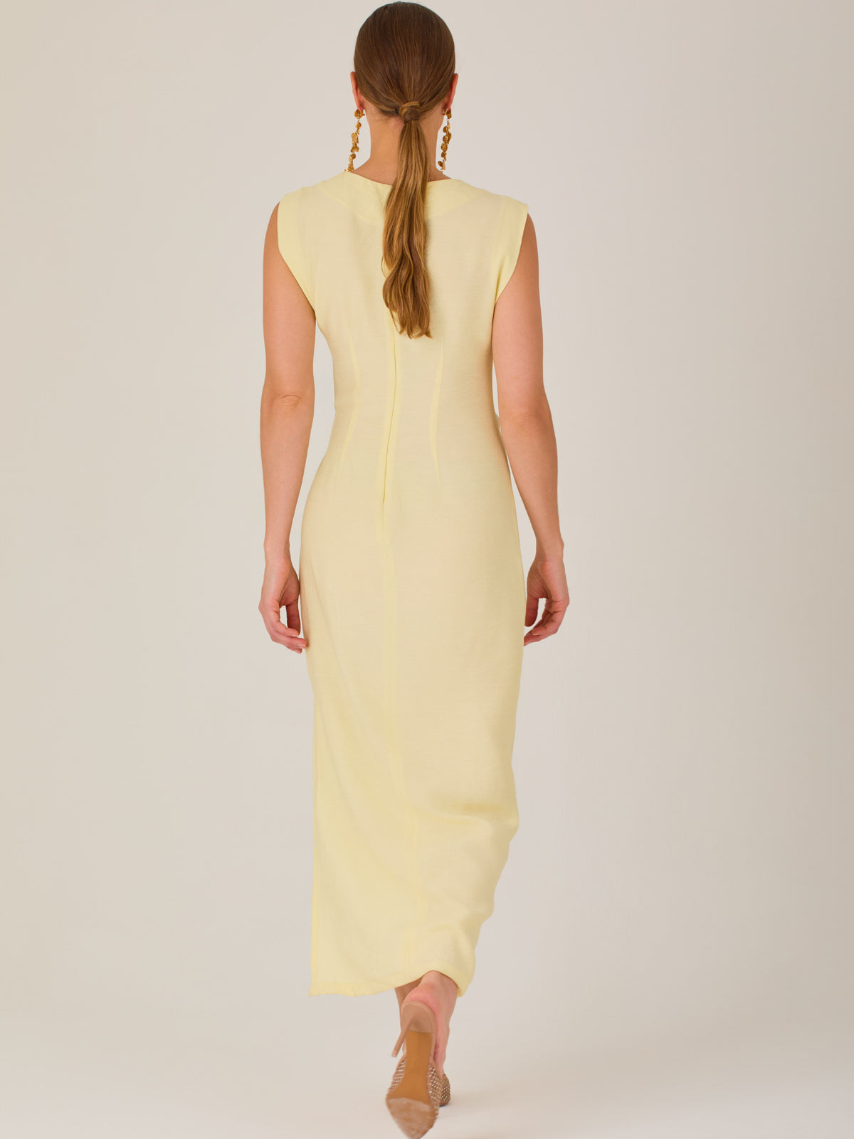 Este vestido es largo y sin mangas, con un diseño elegante y minimalista. Tiene un escote cerrado redondo y un detalle drapeado en la parte frontal del torso, que crea una ligera asimetría visual y acentúa la silueta. Este drapeado se sostiene con una hebilla decorativa al costado de la cintura, lo que añade un toque sofisticado y estilizado. La falda cae de manera recta hasta los tobillos, con una pequeña abertura lateral que proporciona comodidad y un leve movimiento al caminar. El corte general es limpio