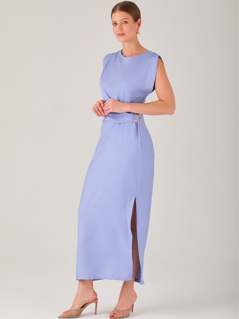 Este vestido es largo y sin mangas, con un diseño elegante y minimalista. Tiene un escote cerrado redondo y un detalle drapeado en la parte frontal del torso, que crea una ligera asimetría visual y acentúa la silueta. Este drapeado se sostiene con una hebilla decorativa al costado de la cintura, lo que añade un toque sofisticado y estilizado. La falda cae de manera recta hasta los tobillos, con una pequeña abertura lateral que proporciona comodidad y un leve movimiento al caminar. El corte general es limpio