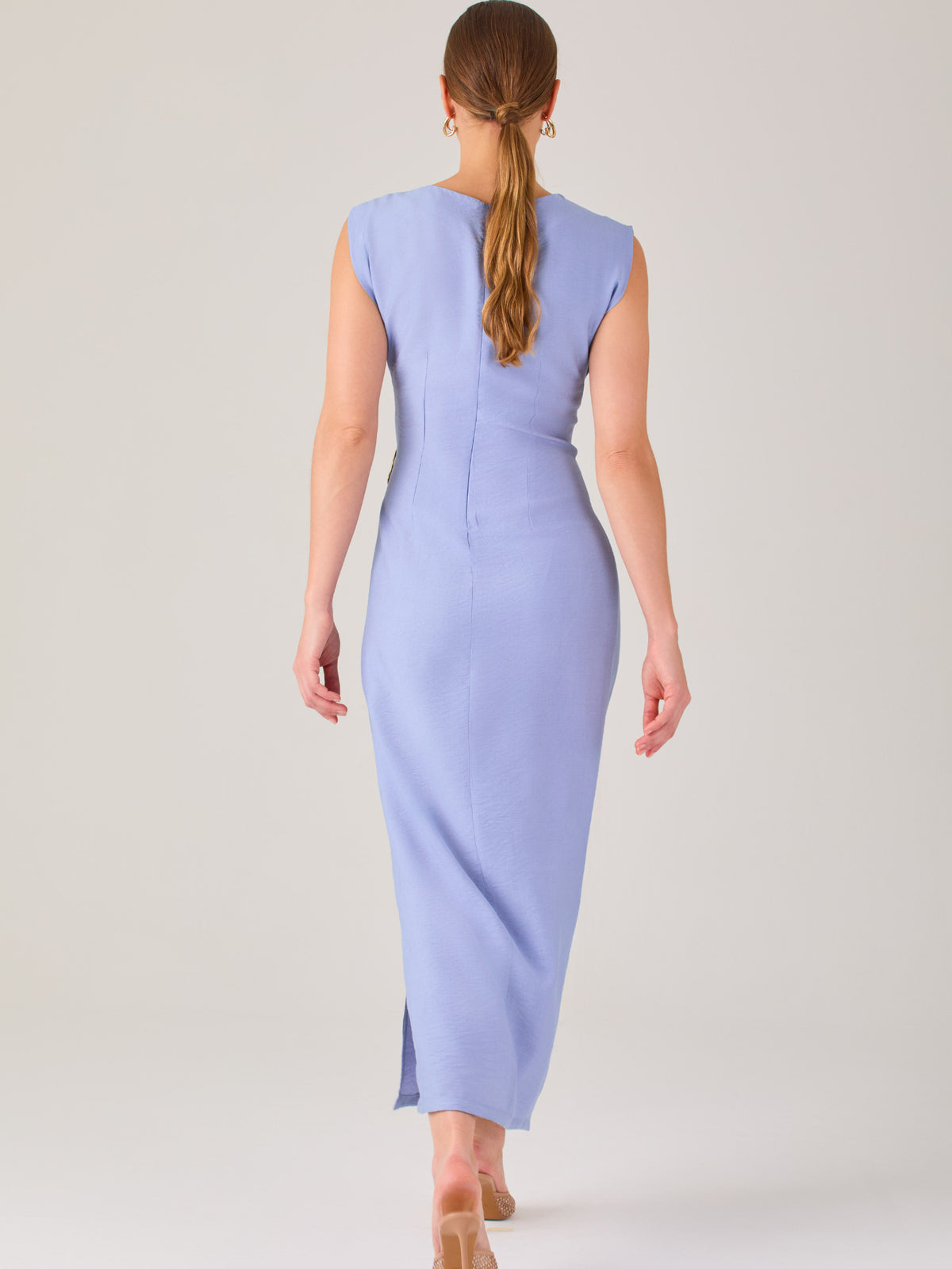 Este vestido es largo y sin mangas, con un diseño elegante y minimalista. Tiene un escote cerrado redondo y un detalle drapeado en la parte frontal del torso, que crea una ligera asimetría visual y acentúa la silueta. Este drapeado se sostiene con una hebilla decorativa al costado de la cintura, lo que añade un toque sofisticado y estilizado. La falda cae de manera recta hasta los tobillos, con una pequeña abertura lateral que proporciona comodidad y un leve movimiento al caminar. El corte general es limpio