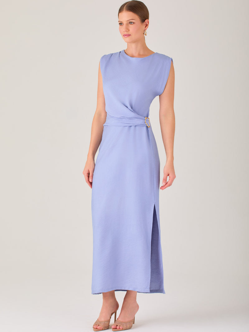 Este vestido es largo y sin mangas, con un diseño elegante y minimalista. Tiene un escote cerrado redondo y un detalle drapeado en la parte frontal del torso, que crea una ligera asimetría visual y acentúa la silueta. Este drapeado se sostiene con una hebilla decorativa al costado de la cintura, lo que añade un toque sofisticado y estilizado. La falda cae de manera recta hasta los tobillos, con una pequeña abertura lateral que proporciona comodidad y un leve movimiento al caminar. El corte general es limpio