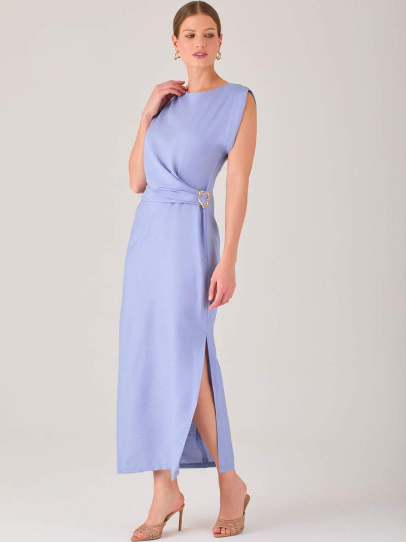Este vestido es largo y sin mangas, con un diseño elegante y minimalista. Tiene un escote cerrado redondo y un detalle drapeado en la parte frontal del torso, que crea una ligera asimetría visual y acentúa la silueta. Este drapeado se sostiene con una hebilla decorativa al costado de la cintura, lo que añade un toque sofisticado y estilizado. La falda cae de manera recta hasta los tobillos, con una pequeña abertura lateral que proporciona comodidad y un leve movimiento al caminar. El corte general es limpio