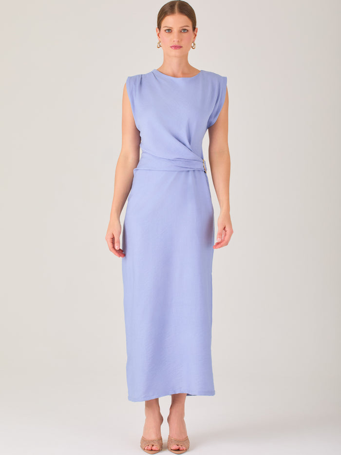 Este vestido es largo y sin mangas, con un diseño elegante y minimalista. Tiene un escote cerrado redondo y un detalle drapeado en la parte frontal del torso, que crea una ligera asimetría visual y acentúa la silueta. Este drapeado se sostiene con una hebilla decorativa al costado de la cintura, lo que añade un toque sofisticado y estilizado. La falda cae de manera recta hasta los tobillos, con una pequeña abertura lateral que proporciona comodidad y un leve movimiento al caminar. El corte general es limpio