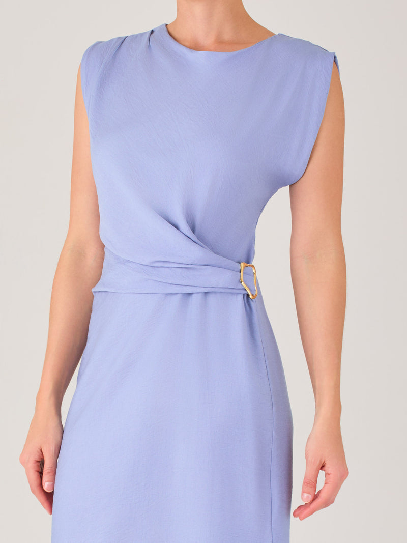 Este vestido es largo y sin mangas, con un diseño elegante y minimalista. Tiene un escote cerrado redondo y un detalle drapeado en la parte frontal del torso, que crea una ligera asimetría visual y acentúa la silueta. Este drapeado se sostiene con una hebilla decorativa al costado de la cintura, lo que añade un toque sofisticado y estilizado. La falda cae de manera recta hasta los tobillos, con una pequeña abertura lateral que proporciona comodidad y un leve movimiento al caminar. El corte general es limpio