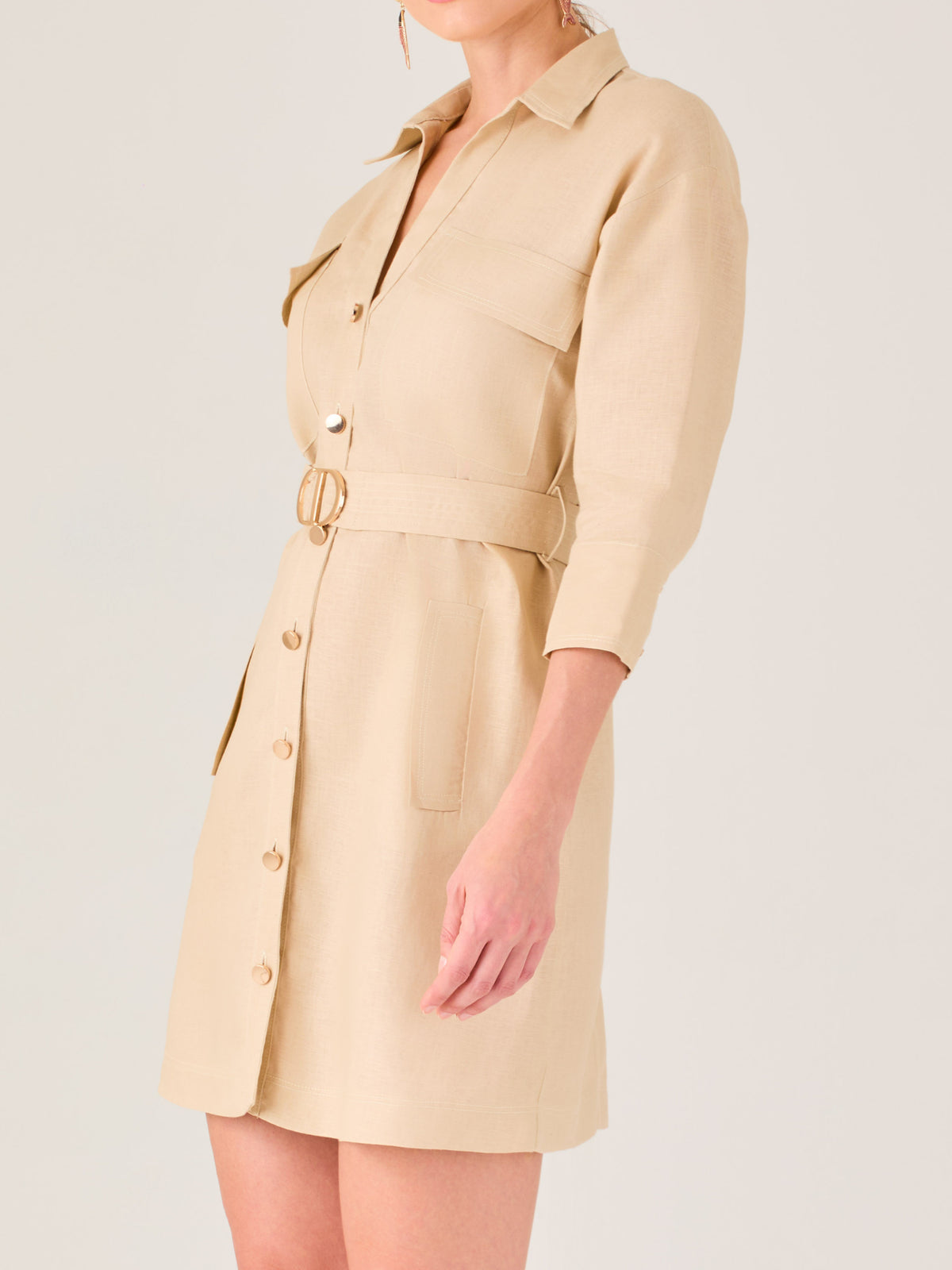 Vestido tipo camisero beige, corto, con cuello clásico, mangas tres cuartos, botones frontales y cinturón con hebilla dorada. Estilo elegante y casual.
