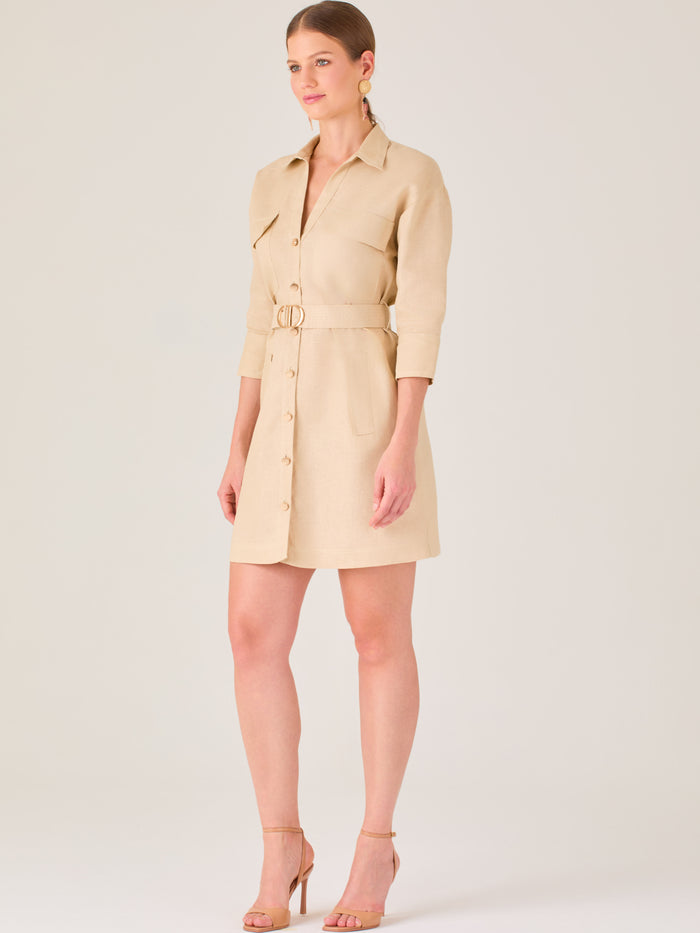 Vestido tipo camisero beige, corto, con cuello clásico, mangas tres cuartos, botones frontales y cinturón con hebilla dorada. Estilo elegante y casual.