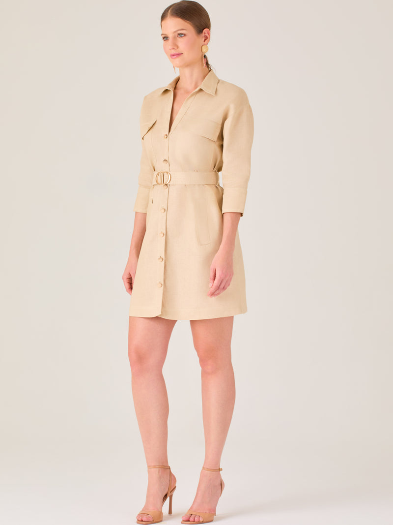 Vestido tipo camisero beige, corto, con cuello clásico, mangas tres cuartos, botones frontales y cinturón con hebilla dorada. Estilo elegante y casual.